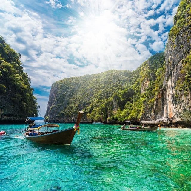 Wooden long-tail boats float on turquoise water in a sunny lagoon surrounded by lush green cliffs.