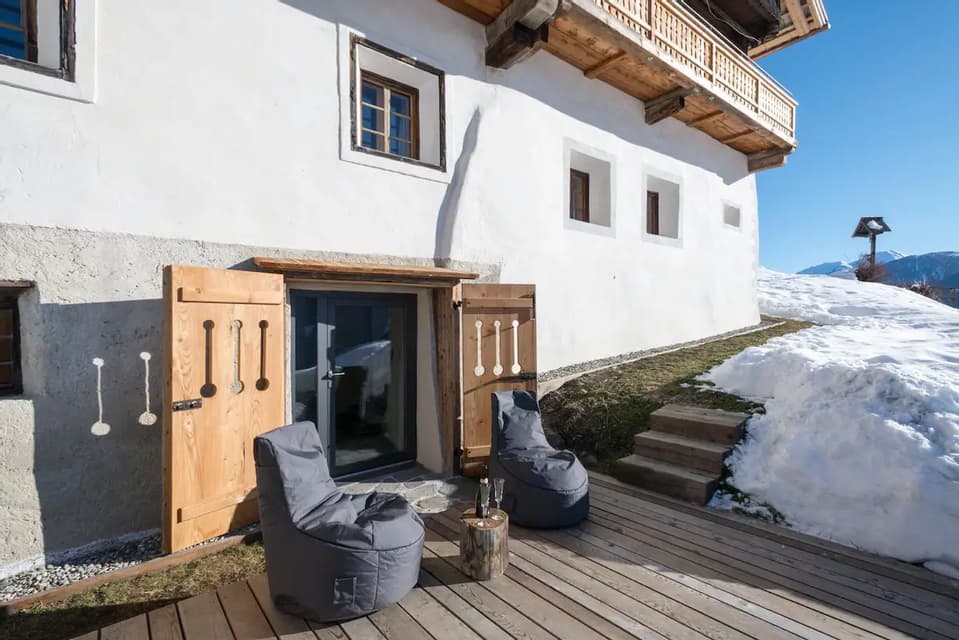 Due poltrone a sacco su una terrazza in legno all'esterno di uno chalet di montagna, con una bottiglia di spumante e neve a terra.