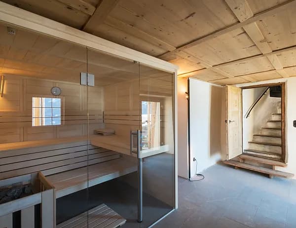 Una sauna moderna con pareti in vetro e panche in legno, situata in una stanza rustica con soffitto in legno e una scala aperta.