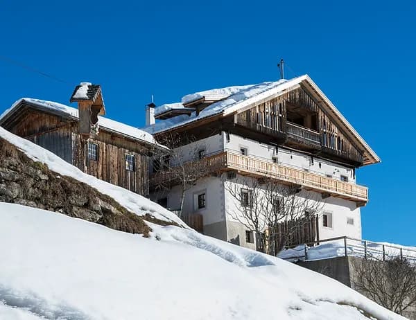 Una tradizionale casa alpina con il tetto coperto di neve si erge su un pendio innevato contro un cielo azzurro limpido.