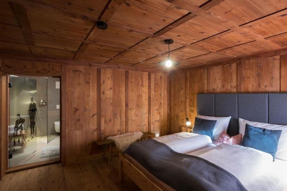 Una camera da letto rustica con pareti e soffitto rivestiti in legno, dotata di un grande letto con testiera grigia e un bagno privato con porta in vetro.