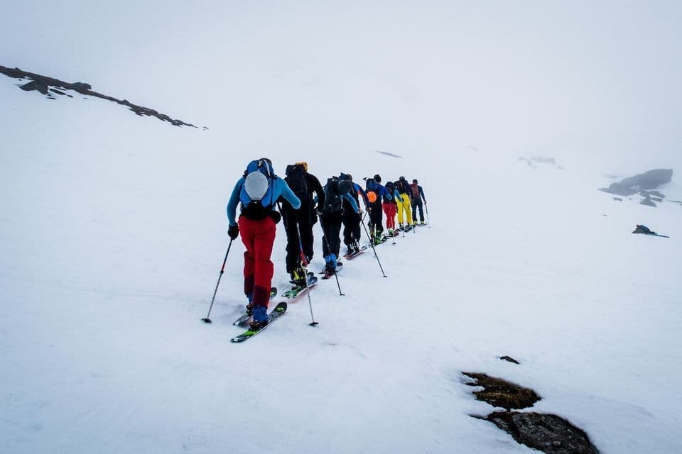 A WeRoad group trip of people ski touring in a line up a vast, snow-covered mountain slope.