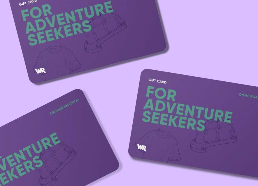 Tre carte regalo WeRoad viola con la scritta 'Per Amanti dell'Avventura' disposte su uno sfondo viola chiaro.