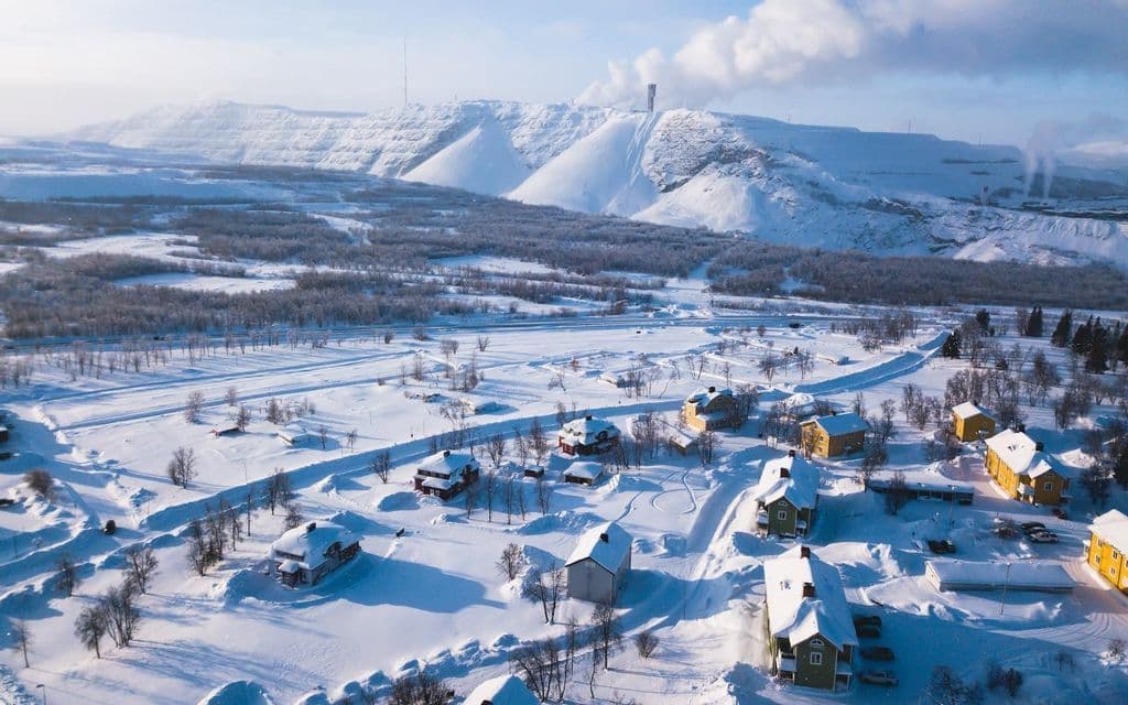 An aerial view of a village with colorful houses covered in deep snow, with a large terraced mountain in the background.