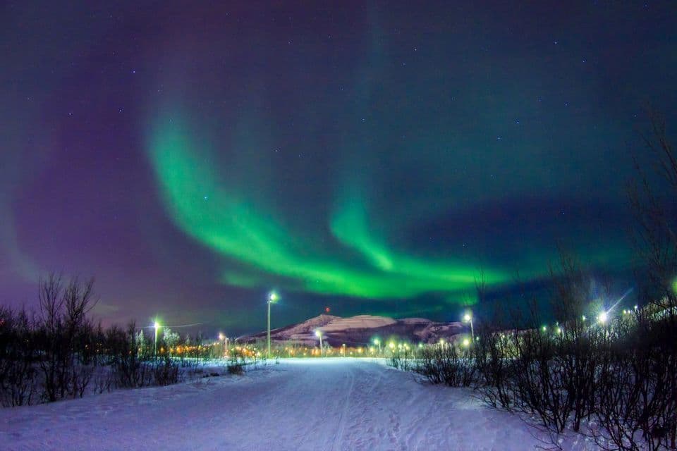 The green Aurora Borealis glows in a starry night sky above a snow-covered landscape with streetlights and a distant mountain.