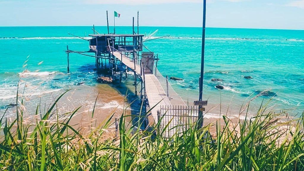 Una capanna da pesca in legno, con una bandiera italiana, sorge su palafitte nel mare turchese ed è collegata alla riva da un lungo pontile.