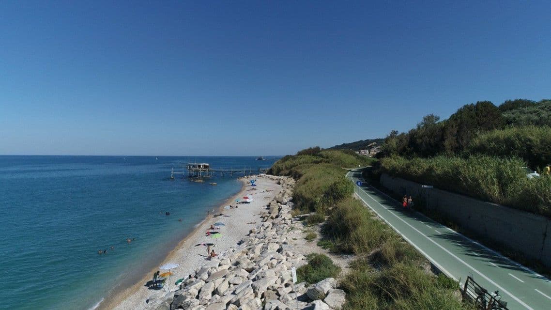 Panoramica aerea di una costa rocciosa, con spiaggia, pista ciclabile verde e un trabucco in legno che si protende nel blu del mare.