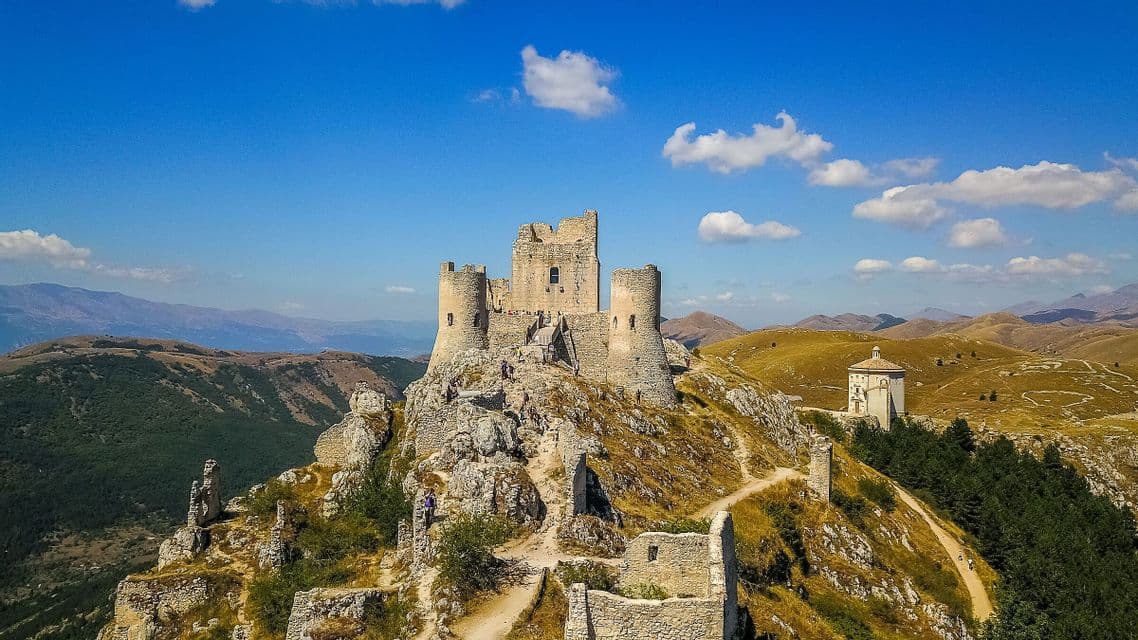 Una vista aerea di un castello in rovina in pietra su una cima montuosa rocciosa, con una piccola cappella e dolci colline sullo sfondo sotto un cielo azzurro.