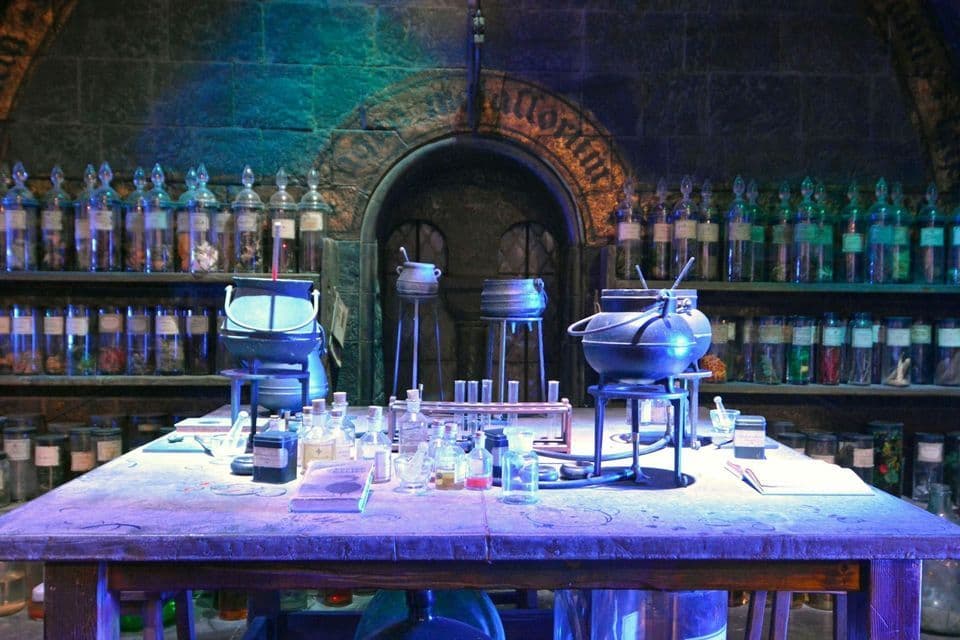 Un'aula di pozioni poco illuminata, con calderoni, fiale e un libro su un tavolo di legno, e scaffali pieni di barattoli di ingredienti lungo una parete di pietra.