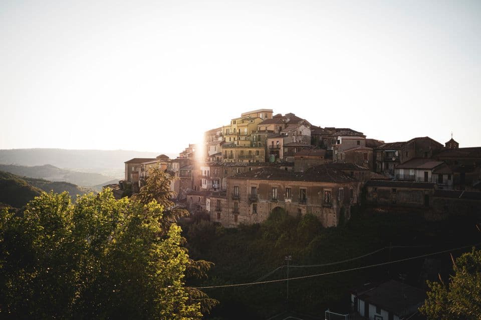 Un borgo di vecchie case costruite su un fianco di collina verdeggiante, con il sole nascente che crea un bagliore luminoso da dietro gli edifici.