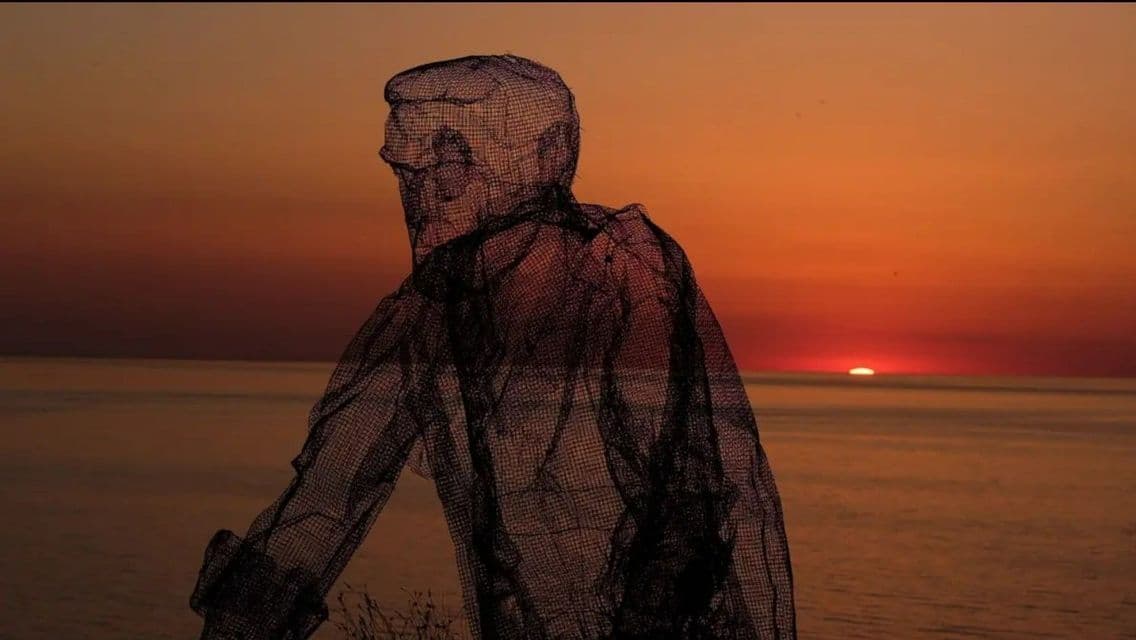 Una scultura in rete metallica traslucida di una persona si staglia in silhouette contro un tramonto arancione sul mare.