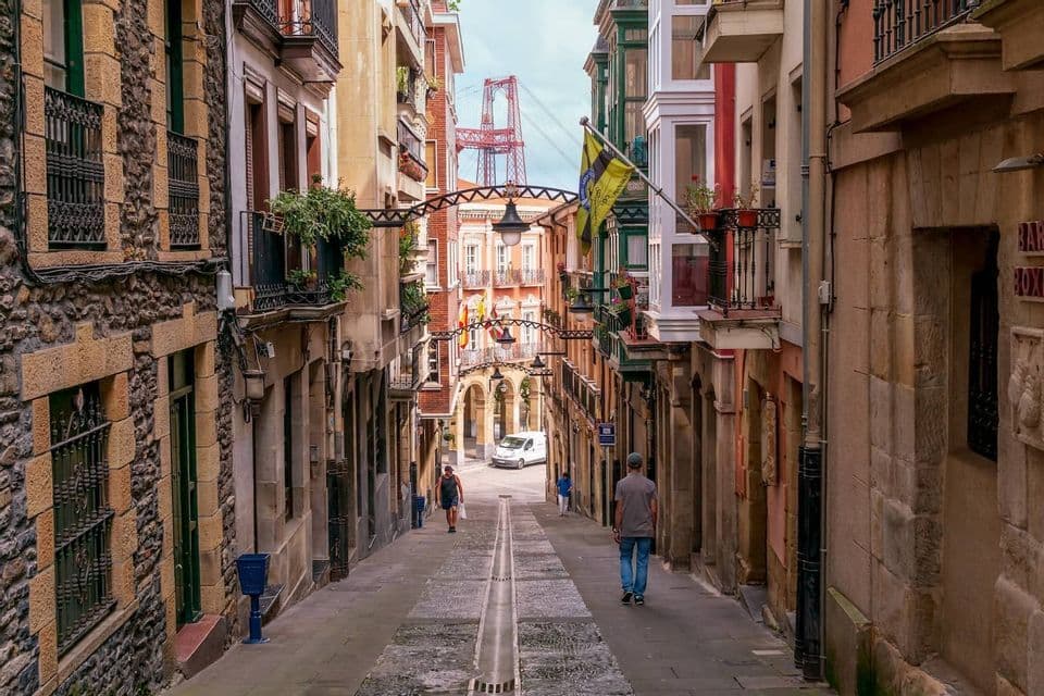 People walk up a narrow, cobblestone street lined with traditional buildings, with a large red bridge visible in the distance.