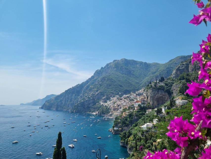 A coastal town built into a steep, green mountainside overlooking a blue bay full of boats, with pink flowers in the foreground.
