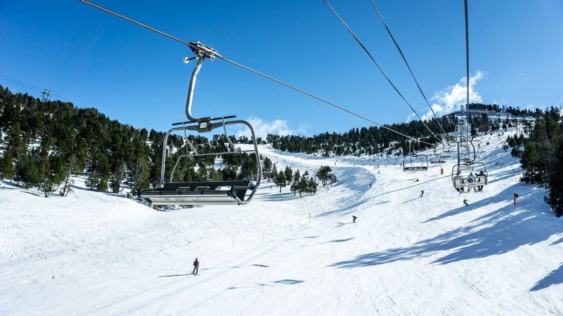 Un telesilla vacío viaja sobre una amplia pista de esquí nevada con esquiadores salpicando la ladera bajo un cielo azul claro.