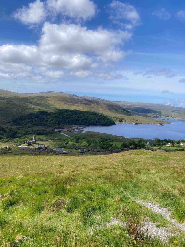 Ein Ausblick von einem grasbewachsenen Hügel über ein Tal mit einer Steinkirche, einem blauen See und sanften Hügeln unter bewölktem Himmel.