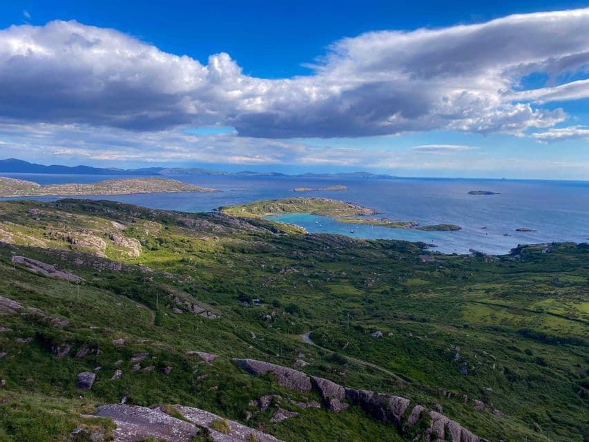 Verdi colline rocciose digradano verso una costa con una baia turchese e diverse piccole isole, sotto un cielo nuvoloso.
