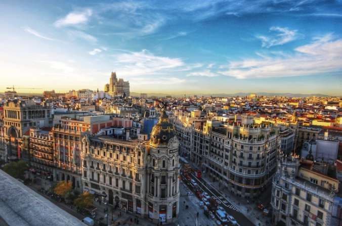 Una vista panoramica di un paesaggio urbano europeo al tramonto, con luce dorata che illumina gli edifici e un cielo blu parzialmente nuvoloso.
