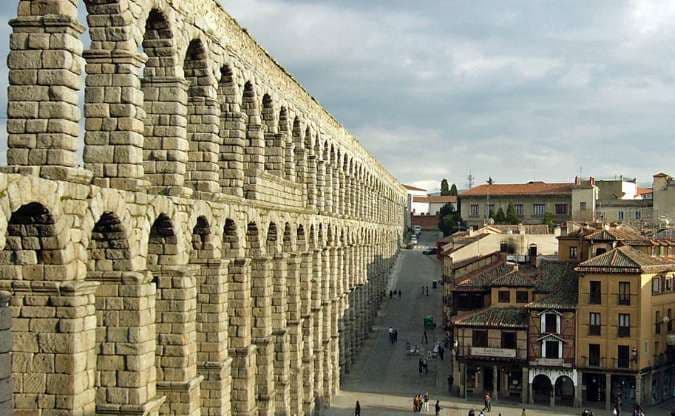 Un antico acquedotto in pietra a due livelli si estende attraverso una storica piazza cittadina con persone che camminano sotto.