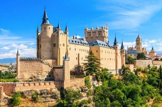 Un grande castello di pietra con molte torrette appuntite si erge su una collina verde e rocciosa sotto un cielo azzurro chiaro.