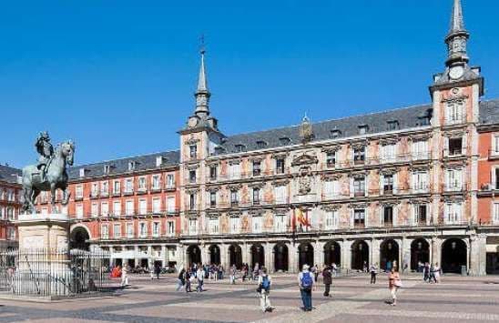 Una statua equestre si erge in un'ampia piazza pavimentata, di fronte a un elaborato edificio storico con guglie, sotto un cielo azzurro e limpido.