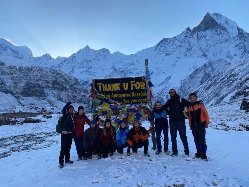 Un viaje en grupo de WeRoad posa para una foto frente al letrero del Campamento Base Annapurna, rodeado de montañas cubiertas de nieve.