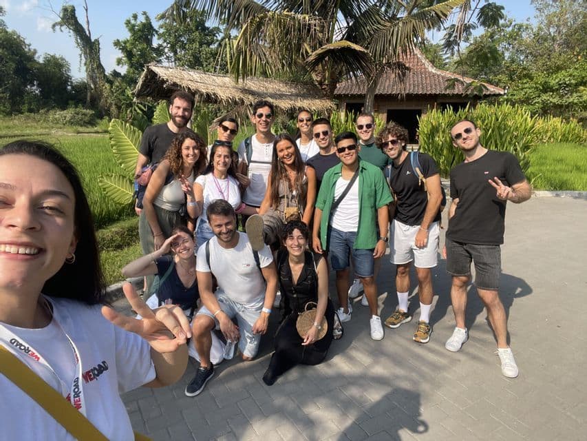 Un groupe WeRoad pose pour un selfie souriant en extérieur, avec des palmiers et une cabane traditionnelle en arrière-plan.