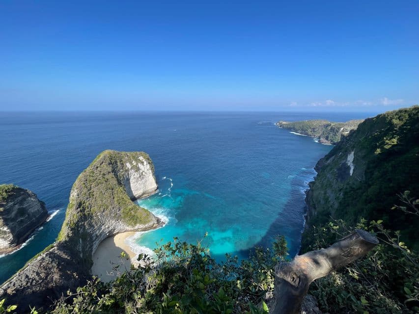 Un mirador elevado con vistas a una costa verde y rocosa, una playa de arena apartada y aguas turquesas bajo un cielo azul claro.