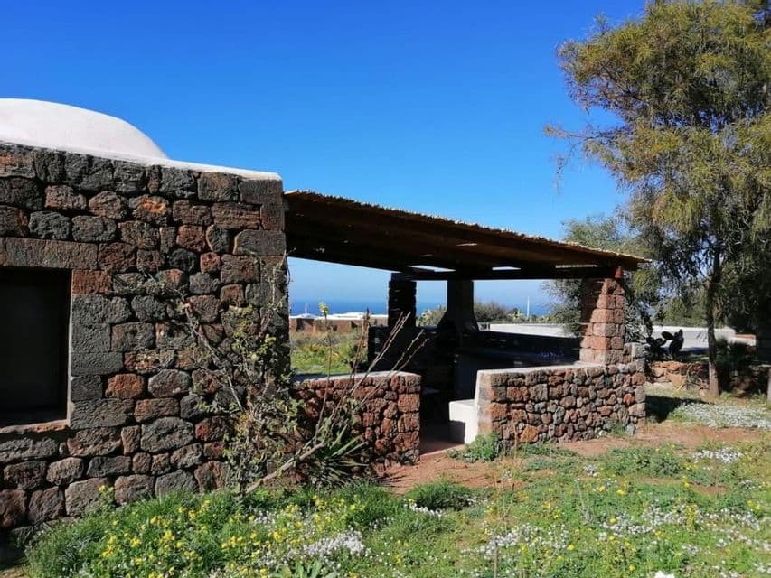 Un edificio rustico in pietra con un patio coperto si erge su una collina erbosa con fiori selvatici, affacciato sul mare sotto un cielo sereno.
