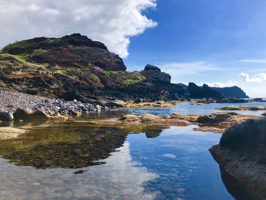 Una tranquilla pozza di marea su una costa rocciosa riflette il cielo blu e le nuvole, con un'auto rossa parcheggiata sulla scogliera sovrastante.