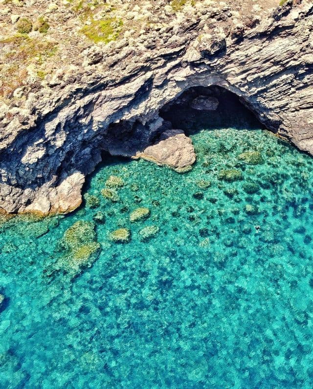 Una vista aerea di una costa rocciosa con una grotta marina che si apre in acque limpide e turchesi con rocce visibili sul fondale.