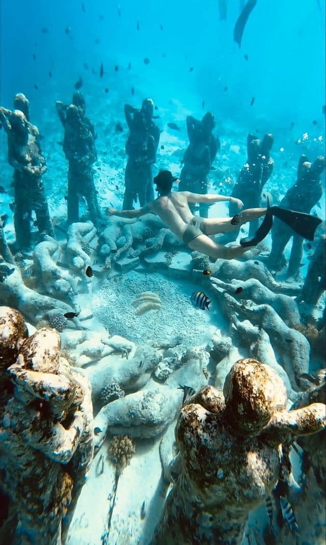 Une personne avec des palmes et un masque fait de la plongée avec tuba au-dessus d'une installation circulaire de statues sous-marines anthropomorphes sur le fond sablonneux de l'océan.