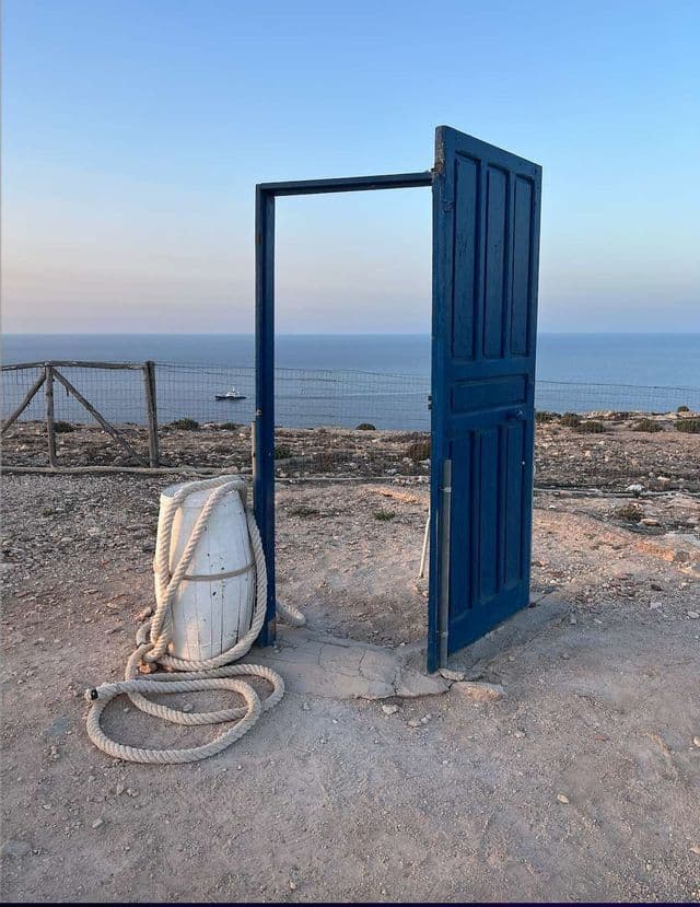 Una porta blu indipendente si apre su una vista del mare da una scogliera al tramonto.