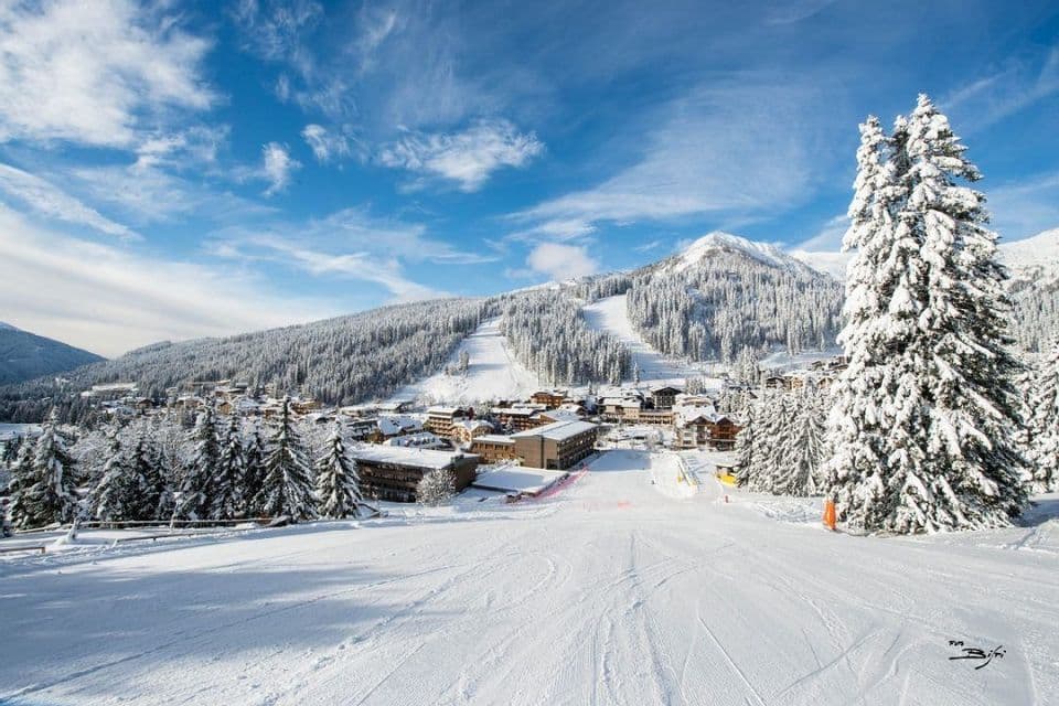 Una pista da sci innevata si affaccia su un villaggio di montagna circondato da foreste di pini sotto un cielo blu con nuvole.