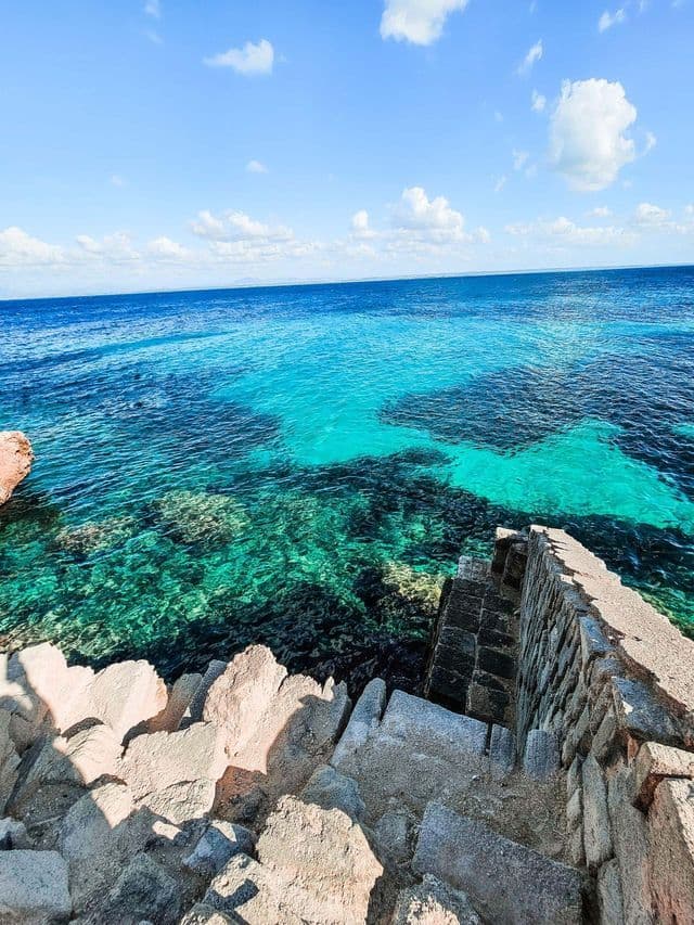 Una vista dall'alto di scale in pietra che scendono verso un mare turchese vibrante lungo una costa rocciosa.