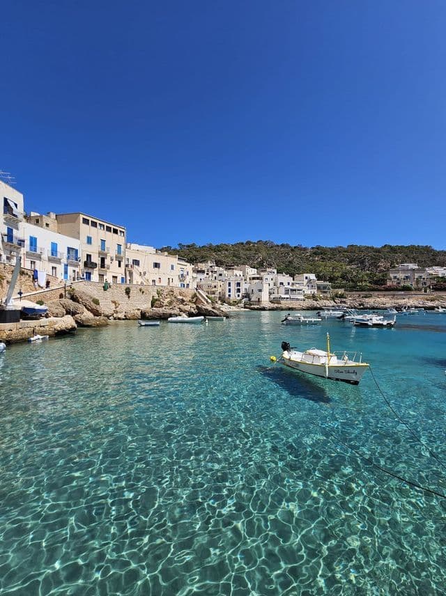 Un villaggio costiero soleggiato con edifici bianchi si affaccia su una baia con acqua cristallina turchese e piccole barche.