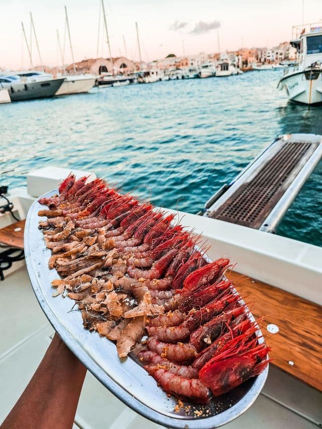 Una mano regge un vassoio d'argento con gamberi rossi freschi e filetti di pesce su una barca in un porto.