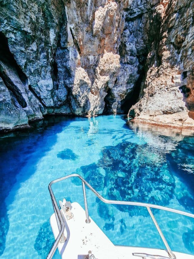Vista dalla prua di una barca che entra in una caletta rocciosa con acqua turchese trasparente.