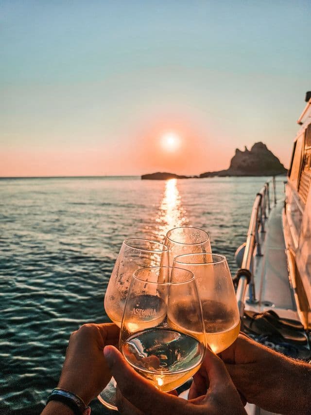 Un viaggio di gruppo WeRoad che brinda con bicchieri di vino su una barca mentre il sole tramonta sull'oceano.
