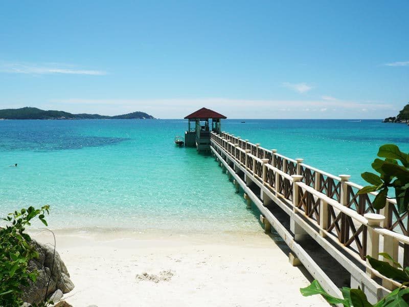 Une jetée en bois s'avance d'une plage de sable blanc dans une eau turquoise claire, avec des îles lointaines sous un ciel bleu éclatant.