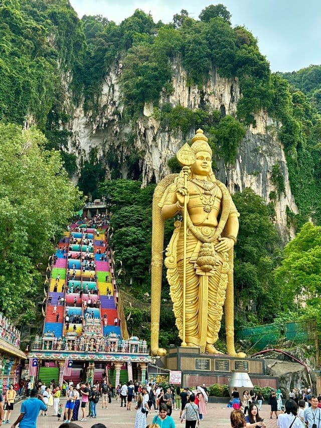 Una gigantesca statua dorata di una divinità si erge accanto a una lunga e colorata scalinata dove le persone salgono su una montagna lussureggiante e verde.