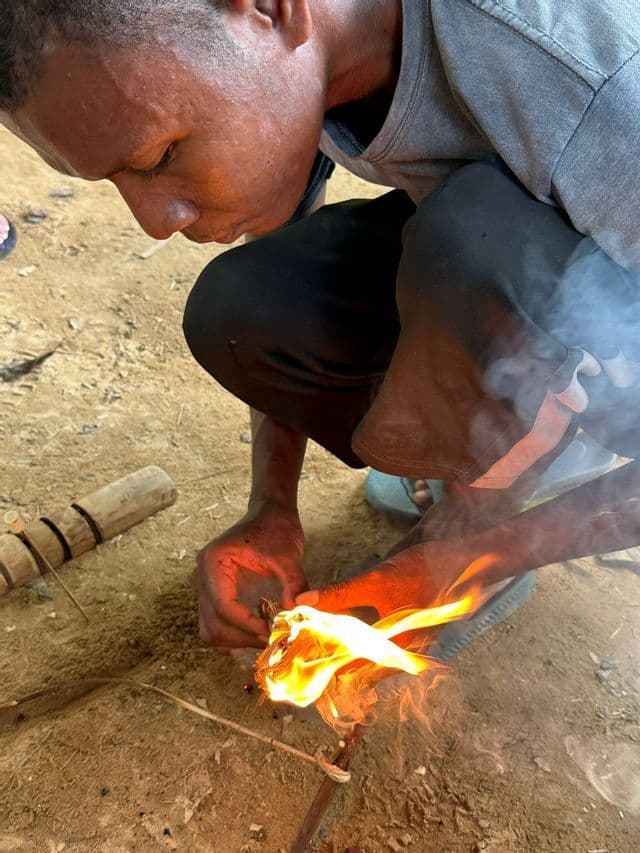 Un uomo si accovaccia per accendere un fuoco a mano, tenendo un fascio di esca fiammeggiante sopra dei bastoncini.