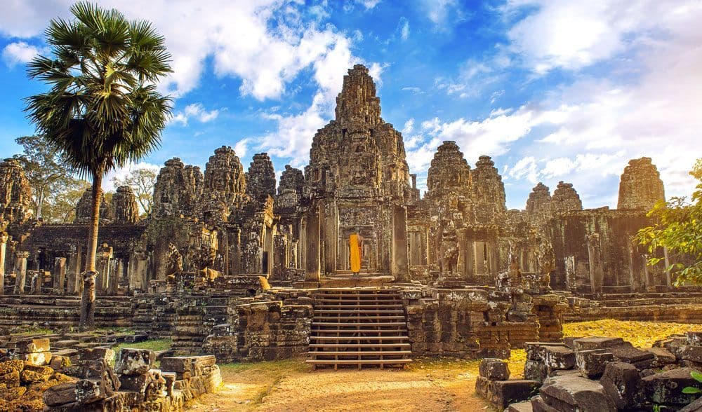 A person in orange robes walks up the stone steps of a large, ancient temple with carved towers, next to a tall palm tree.