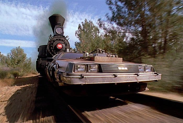Un coche DeLorean modificado es empujado a gran velocidad por una locomotora de vapor a lo largo de una vía férrea a través de una zona boscosa.
