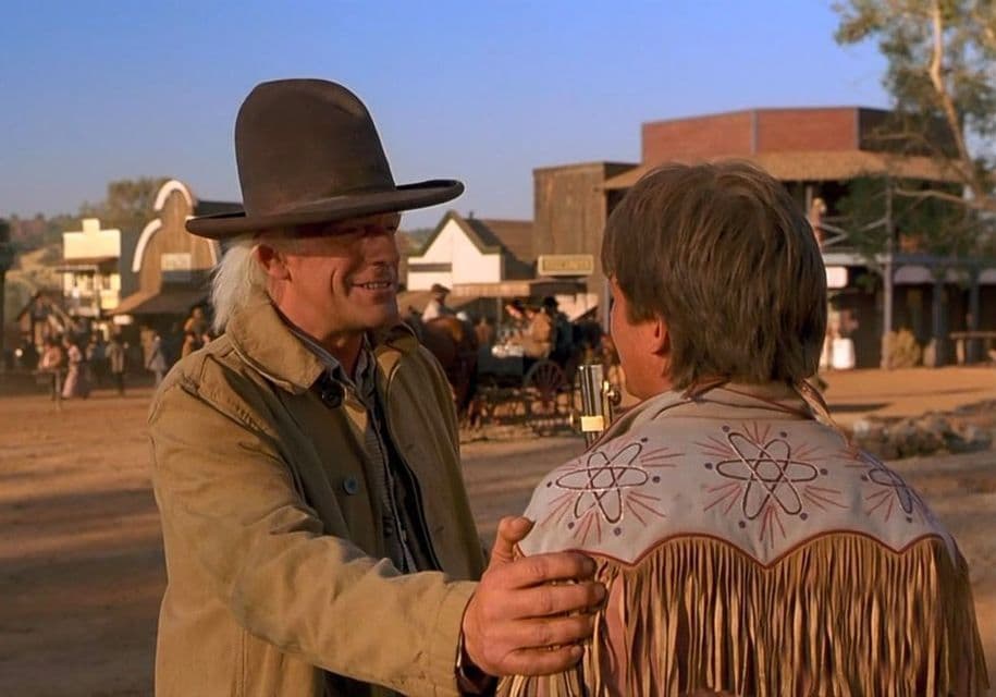 Un uomo con un cappello alto marrone sorride a un altro uomo che indossa una giacca sfrangiata con simboli atomici in una polverosa città del Vecchio West.