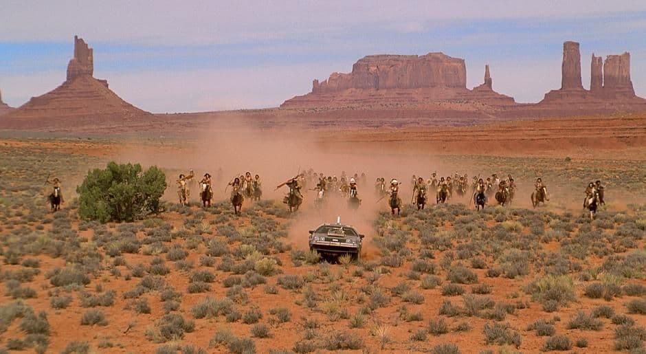 Un grupo de nativos americanos a caballo persigue un coche por un desierto con grandes formaciones rocosas rojas al fondo.