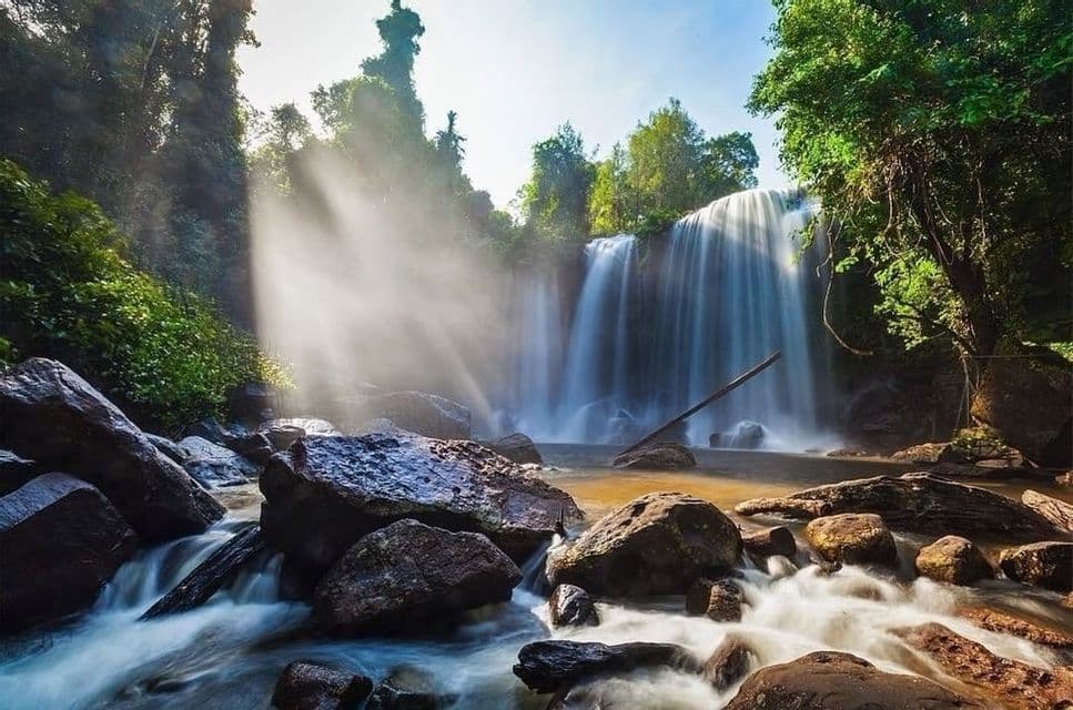 Una amplia cascada cae en un arroyo rocoso en un bosque frondoso, con rayos de sol brillando a través de la niebla.
