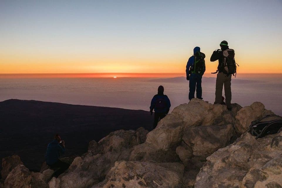 Un viaggio di gruppo WeRoad con zaino in spalla ammira l'alba da una vetta rocciosa sopra un mare di nuvole.