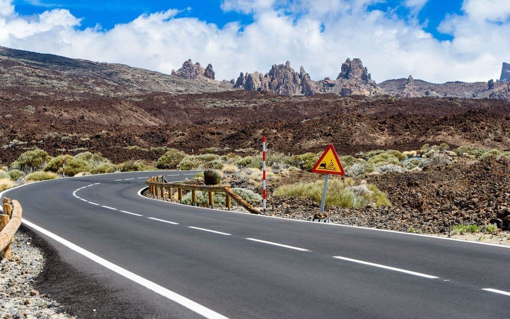 Una strada asfaltata tortuosa attraverso un paesaggio vulcanico con un segnale di 'caduta massi' e montagne frastagliate sullo sfondo.