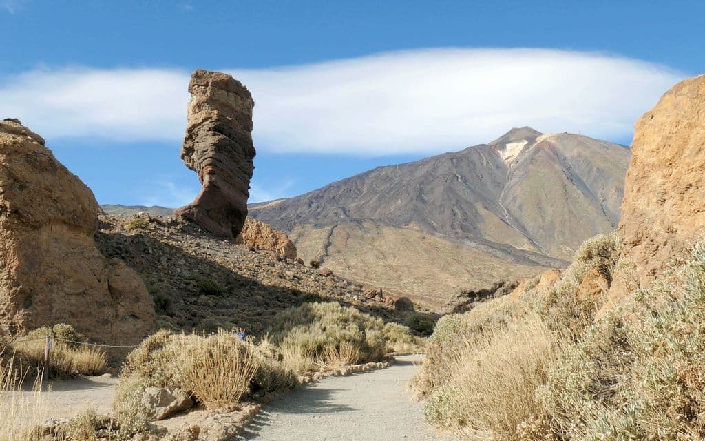 Un sentiero sterrato e tortuoso porta a un'alta formazione rocciosa stratificata in un paesaggio vulcanico, con una montagna sullo sfondo.