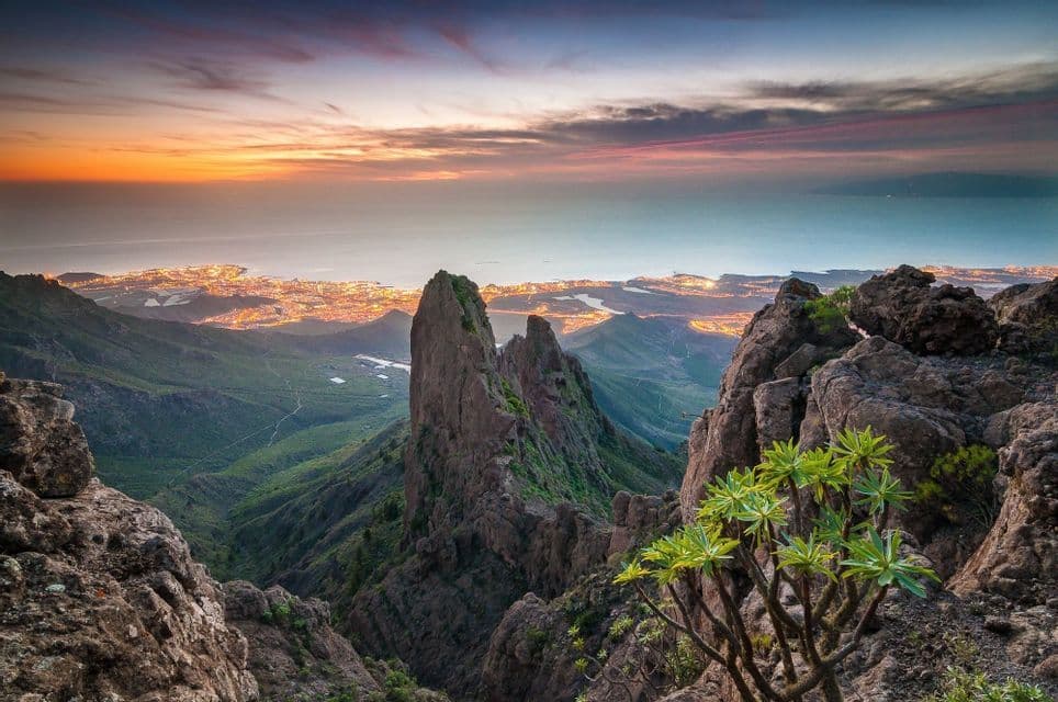 Una vista panoramica da una montagna rocciosa su una valle verde e una città costiera illuminata sul mare al tramonto.